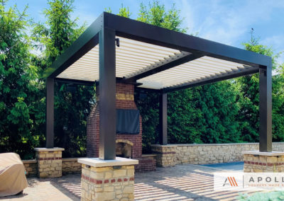 Custom frame configuration of this louvered pergola wraps around fireplace and features custom stonework at post bases.