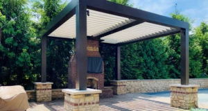 Louvered patio cover shading an outdoor brick fireplace area surrounded by lush greenery
