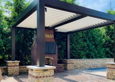 Louvered patio cover shading an outdoor brick fireplace area surrounded by lush greenery