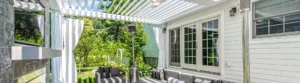 Modern white louvered pergola attached to house with French doors, stone fireplace, and outdoor seating area.