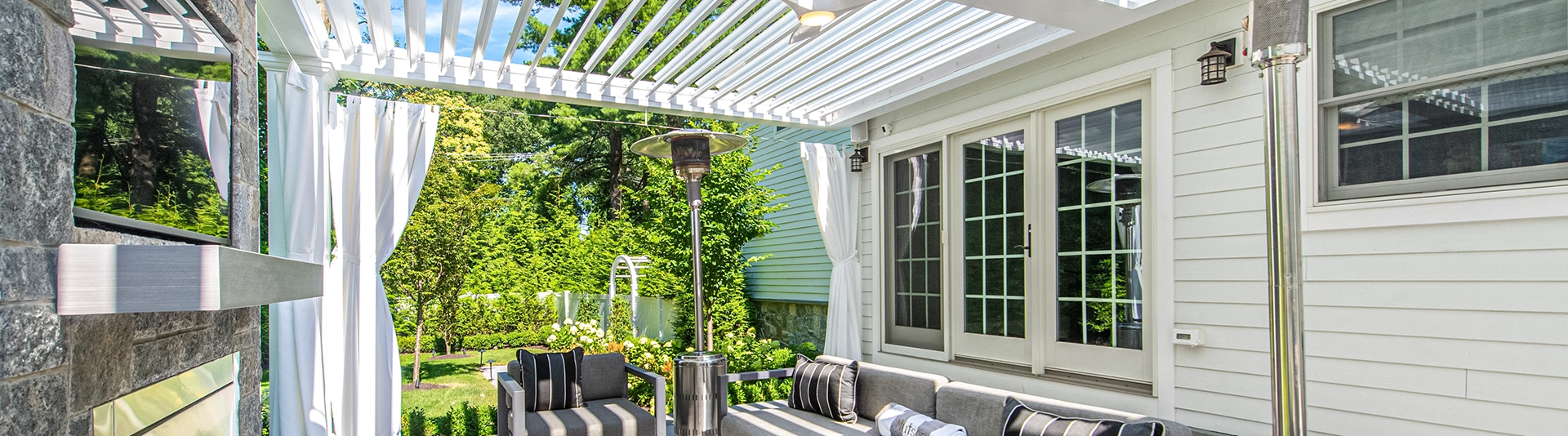Modern white louvered pergola attached to house with French doors, stone fireplace, and outdoor seating area.