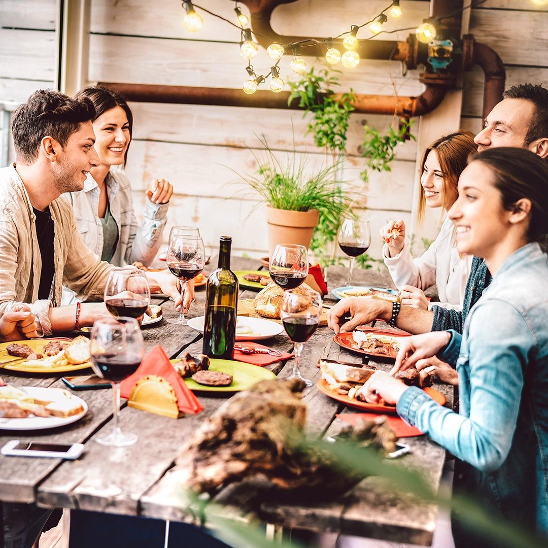 Group of joyful friends enjoying a BBQ and wine outdoors in the shade.</p>
<p>