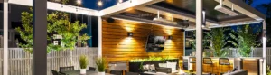 Modern outdoor patio with wooden walls, louvered roof system, hanging string lights, and a flat-screen TV surrounded by lush greenery at night.
