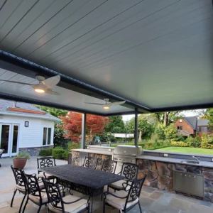 Elegant outdoor dining area under an Apollo louvered pergola, featuring ceiling fans, a stone countertop with built-in grill, and lush garden surroundings.