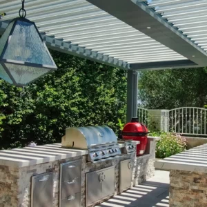 Modern outdoor kitchen beneath a louvered pergola featuring a distinctive geometric light fixture, stainless steel appliances, and a vibrant red grill.