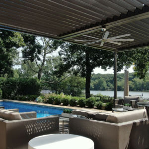 Luxury backyard patio with louvered pergola overlooking a serene lake, complete with outdoor furniture and pool.