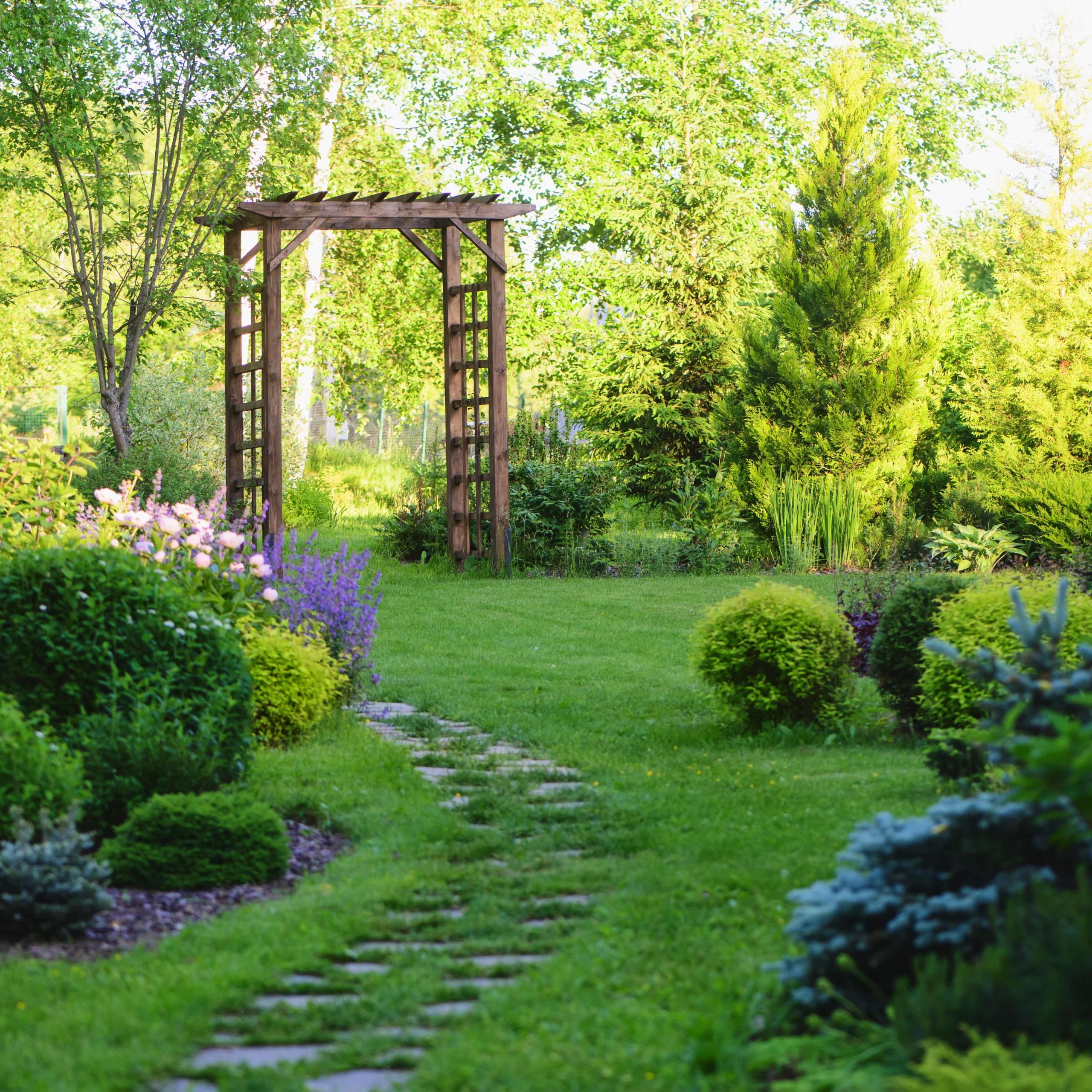 Rustic wooden arbor over a garden path surrounded by lush greenery and blooming flowers.