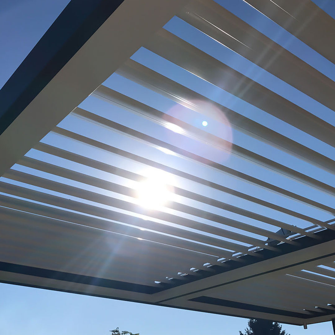 Sunlight filtering through the slats of an open louvered pergola system against a clear blue sky.