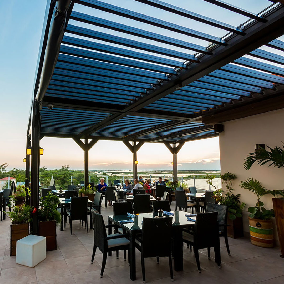 Waterfront restaurant dining under a blue louvered pergola at sunset.