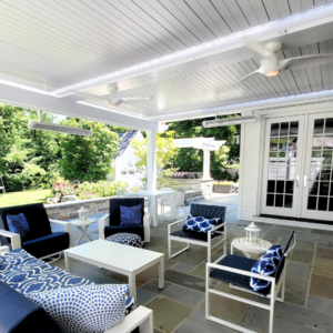 Comfortable outdoor lounge under a white louvered pergola with fans and heaters, adjacent to a home with open French doors.