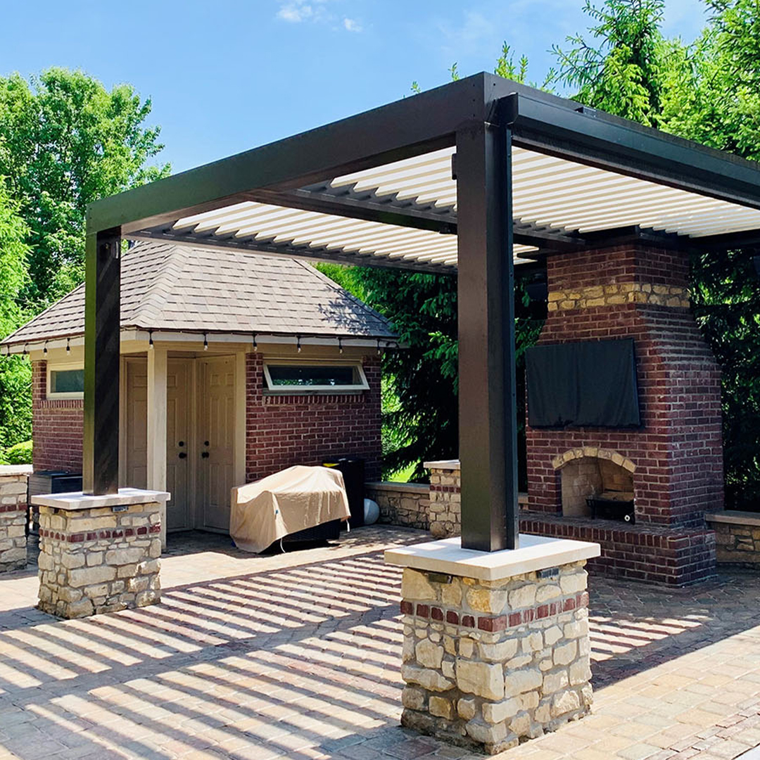 Louvered pergola casting stripes on stone patio with brick fireplace