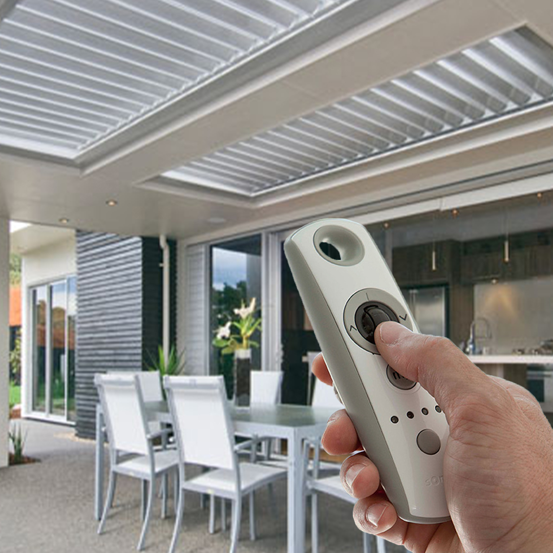 Hand holding remote to adjust motorized louvered roof over modern patio