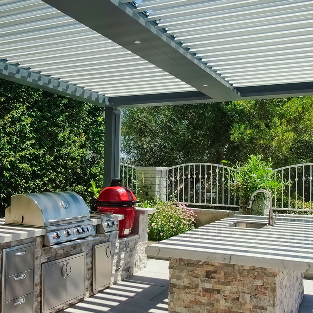 Outdoor kitchen with grill beneath a louvered pergola roof