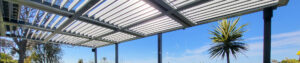Panoramic view of a wall-attached louvered pergola with open louvers, slim support posts, blue sky, and landscaped outdoor setting.