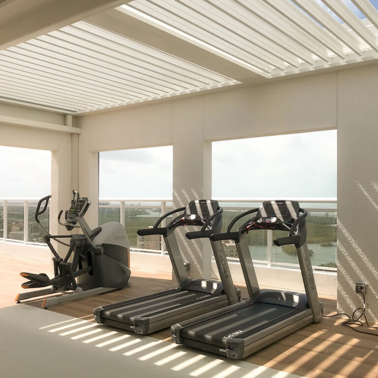 White motorized louvered pergola partially open above a rooftop gym with treadmills and an elliptical, casting striped light across the floor with a coastal city view