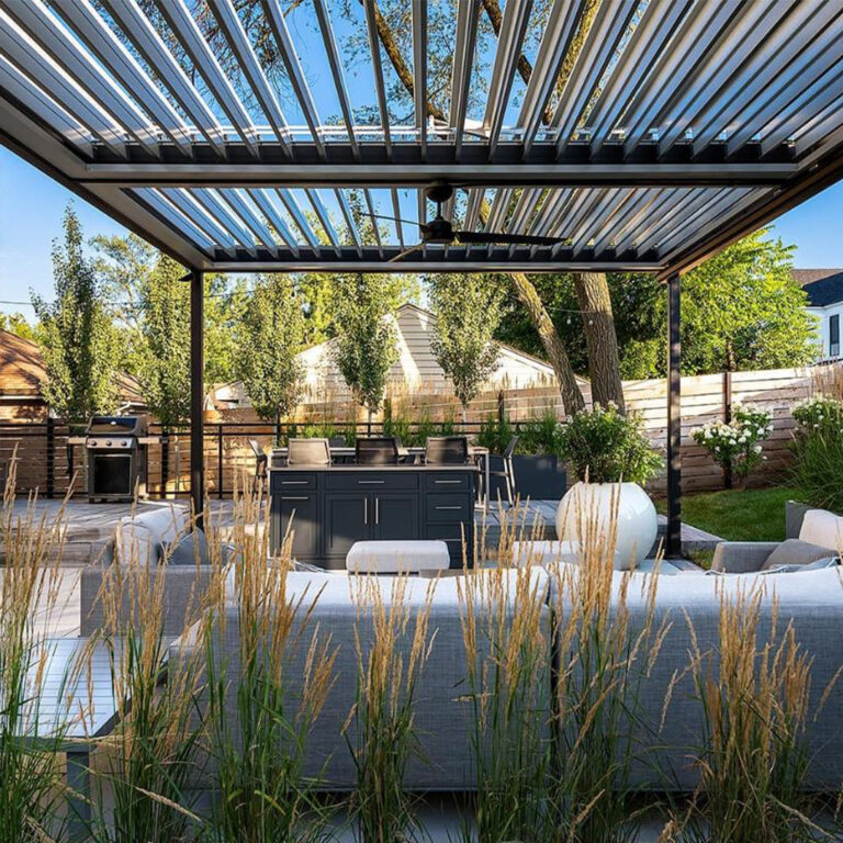 Dark-framed aluminum louvered pergola with open louvers and ceiling fan over a backyard outdoor kitchen, lounge seating, and ornamental grasses in a landscaped yard