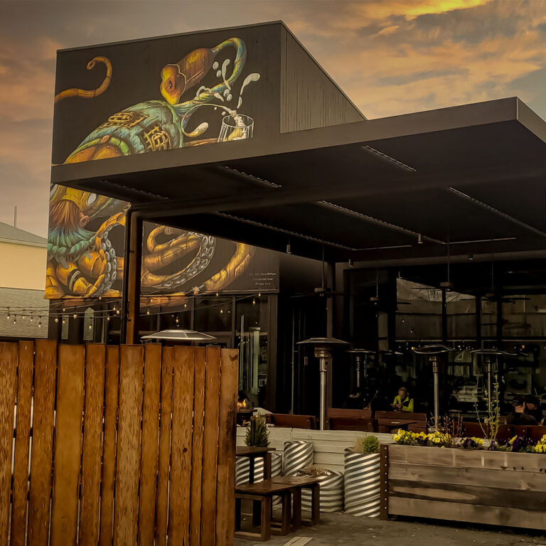 Closed aluminum louvered pergola covering a brewery patio with standing heaters, string lights, and guests seated beneath a large mural at sunset