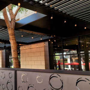 Dark aluminum louvered pergola with partially open louvers and warm string lights over a commercial restaurant patio at dusk, with a mature tree growing through the structure