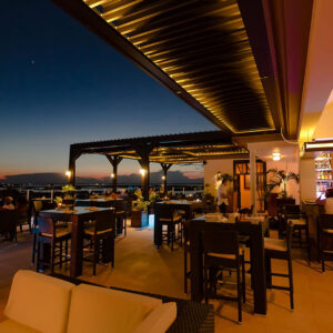 Closed aluminum louvered roof with built-in LED lighting covering a rooftop restaurant terrace at dusk, guests dining with a waterfront horizon in the background