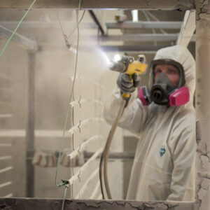 A masked and suited technician powder coating aluminum louver slats hanging on racks inside an industrial spray booth, with pigment misting the air