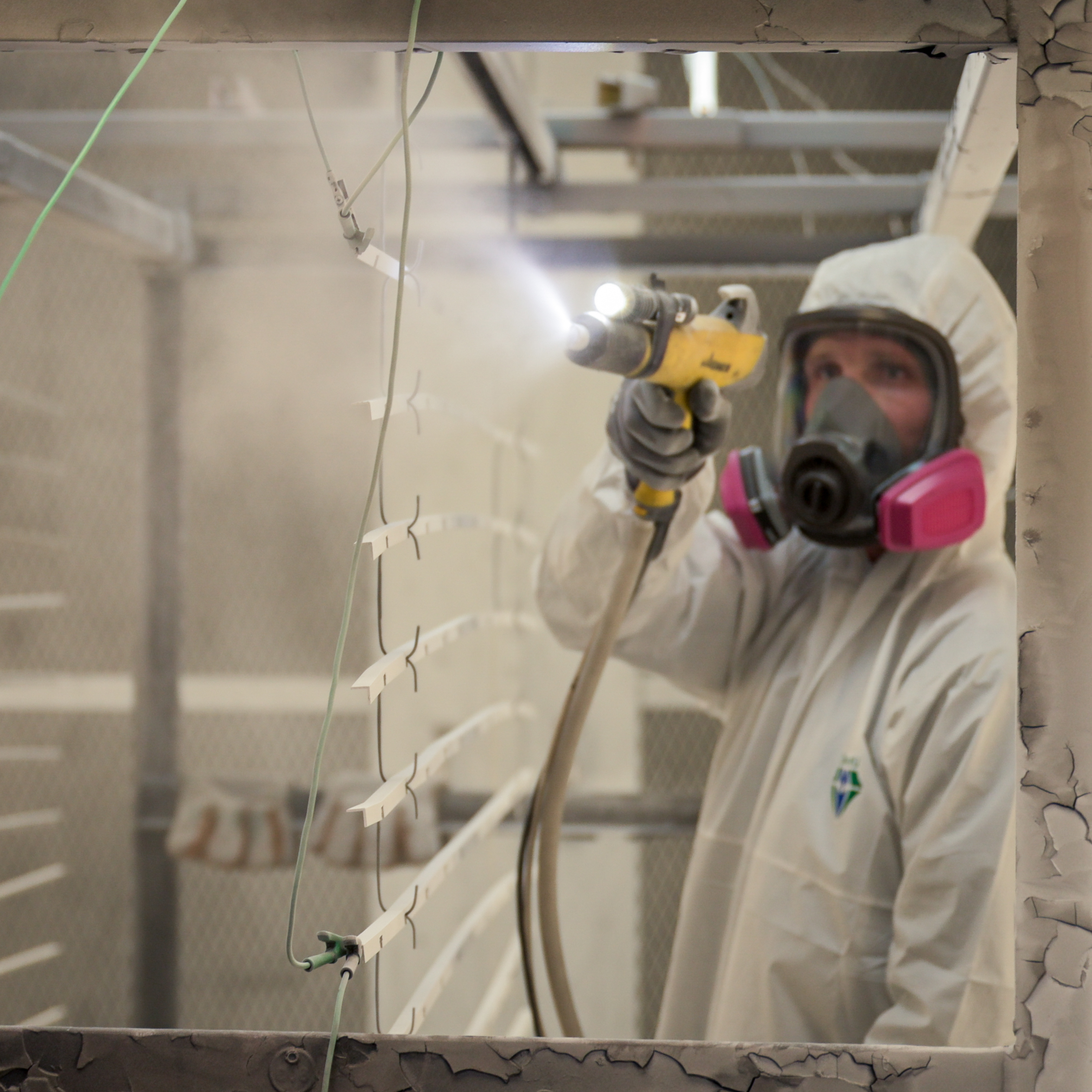 A masked and suited technician powder coating aluminum louver slats hanging on racks inside an industrial spray booth, with pigment misting the air