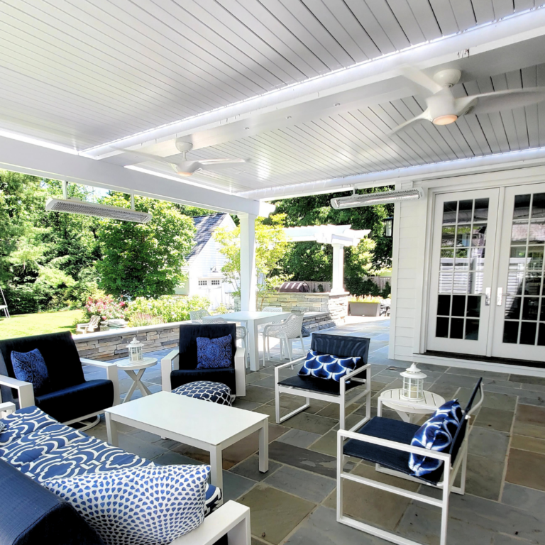 White louvered pergola closed overhead on a stone patio, with two ceiling fans, infrared heaters mounted to the frame, and blue and white outdoor lounge furniture