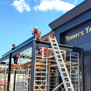 Two installers on scaffolding assembling the aluminum frame of a commercial louvered pergola outside a restaurant storefront