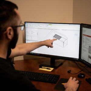 An Apollo designer pointing at a 3D louvered pergola model on a dual-monitor Revit workstation, with project information visible on the second screen.