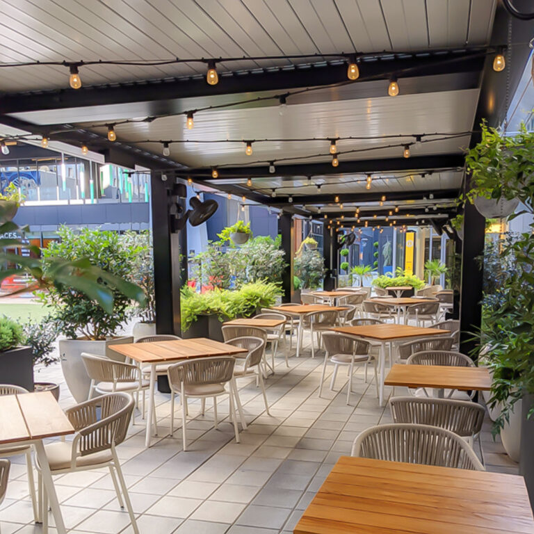 Closed aluminum louvered roof covering a commercial restaurant terrace with string lights, dining tables, and lush planters along an urban streetfront