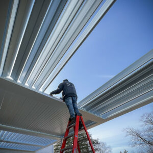 Authorized Apollo dealer installer on a ladder assembling white aluminum louvered pergola louvers against a clear blue sky