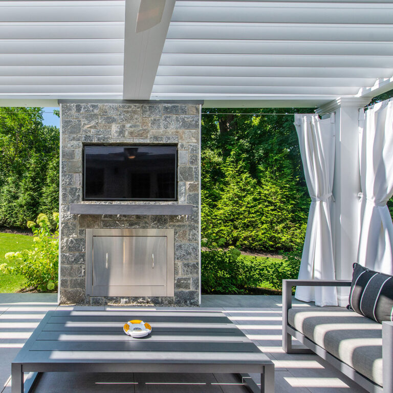 Stone fireplace wall with TV beneath white pergola and curtains in Tenafly NJ