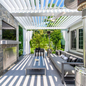Covered outdoor lounge with stone fireplace under white louvered pergola in Tenafly NJ