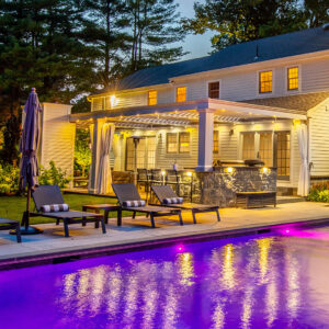 Poolside view of illuminated white louvered pergola and patio at night in Tenafly NJ