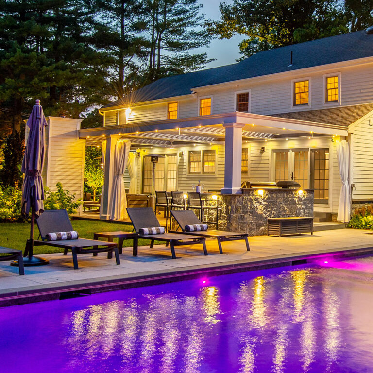 Poolside view of illuminated white louvered pergola and patio at night in Tenafly NJ