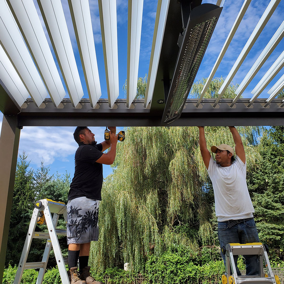 Two installers on ladders mounting an infrared heater to the beam of an open aluminum louvered pergola in a landscaped backyard