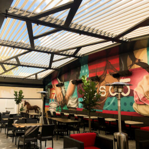 Open aluminum louvered pergola with standing heaters and track lighting over a restaurant dining patio with a large colorful mural on the back wall