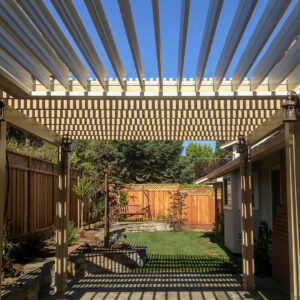 Cream aluminum louvered pergola with louvers partially open casting a checkerboard shadow pattern across a stucco wall and flagstone patio, with lantern post lights and a landscaped backyard beyond