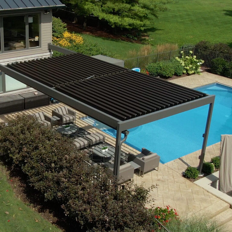 Aerial view of a taupe aluminum louvered pergola with closed dark louvers attached to a contemporary home, covering a striped lounge seating area on a paver patio directly beside a large rectangular pool