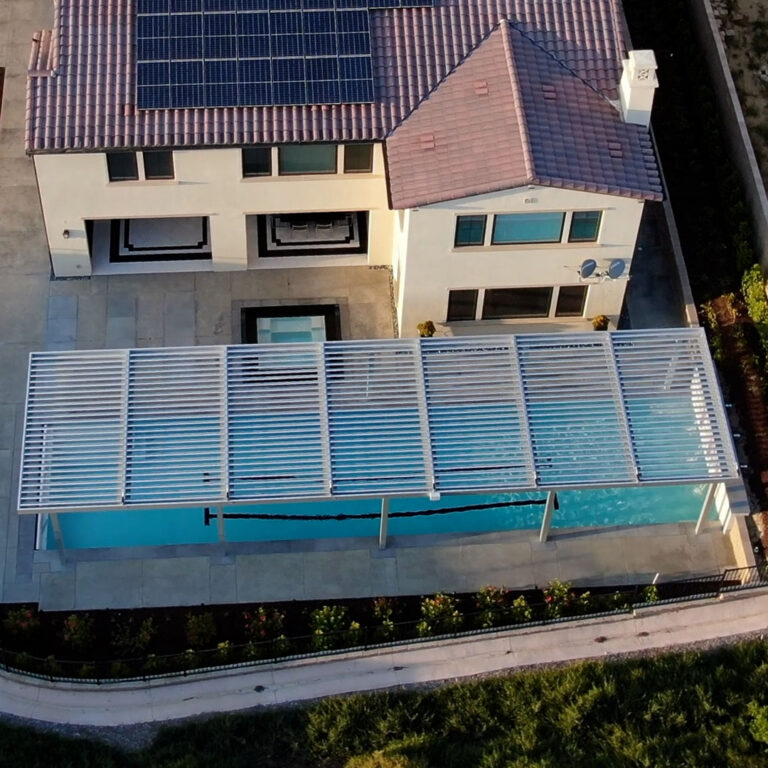Overhead drone shot of a wide white aluminum louvered pergola spanning the full length of a residential pool beside a two-story stucco home