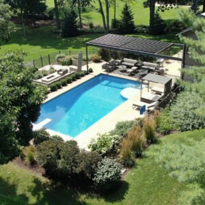 Aerial view of a dark aluminum louvered pergola with closed louvers over a poolside lounge beside a large rectangular pool, fire pit, and ornamental landscaping on a wooded estate property