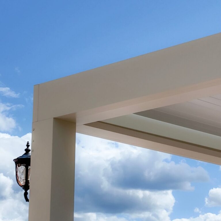 Close-up of a white powder coated aluminum pergola corner joint showing a clean post-to-beam connection with a wall-mounted lantern against a blue sky