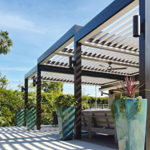 Large dark-framed aluminum pergola with open louvers, a ceiling fan, and column-mounted uplights casting striped shadows across a concrete patio with decorative planters and palm trees