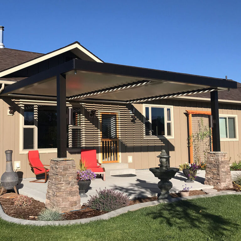 Dark-framed aluminum louvered pergola with partially open louvers casting shadow stripes on a craftsman home, with stone pier bases, red lounge chairs, a chiminea, and a garden fountain on a concrete patio
