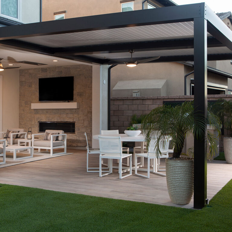 Dark-framed aluminum louvered pergola with closed louvers and dual ceiling fans attached to a stucco home over a patio with dining table, lounge seating, stone fireplace, and outdoor TV at dusk