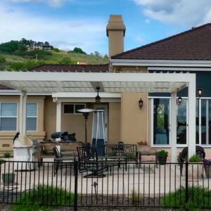 White aluminum louvered pergola attached to a stucco home covering a patio with a tower heater, ceiling fan, dining set, and built-in grill on a hillside property