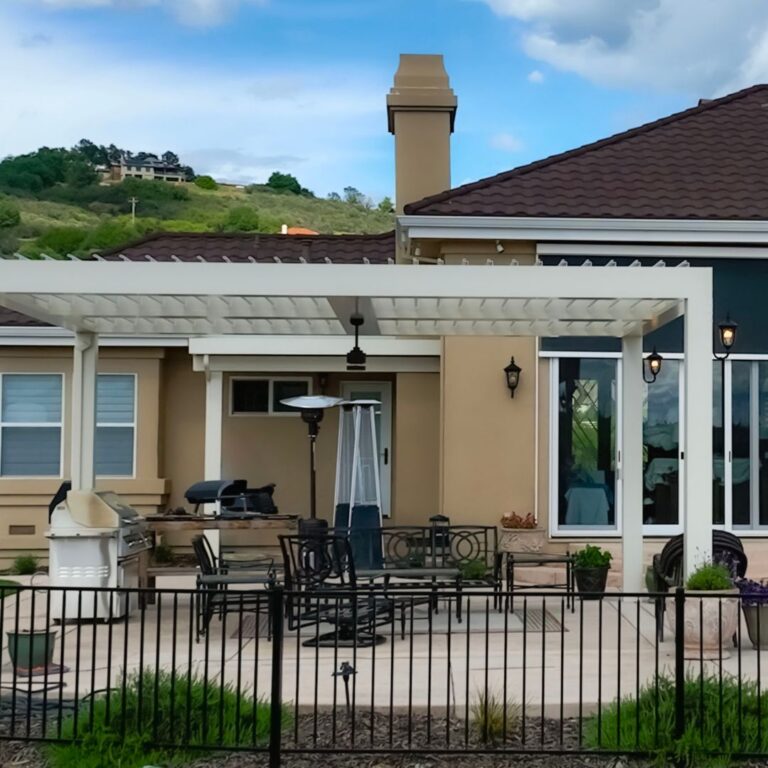 White aluminum louvered pergola attached to a stucco home covering a patio with a tower heater, ceiling fan, dining set, and built-in grill on a hillside property