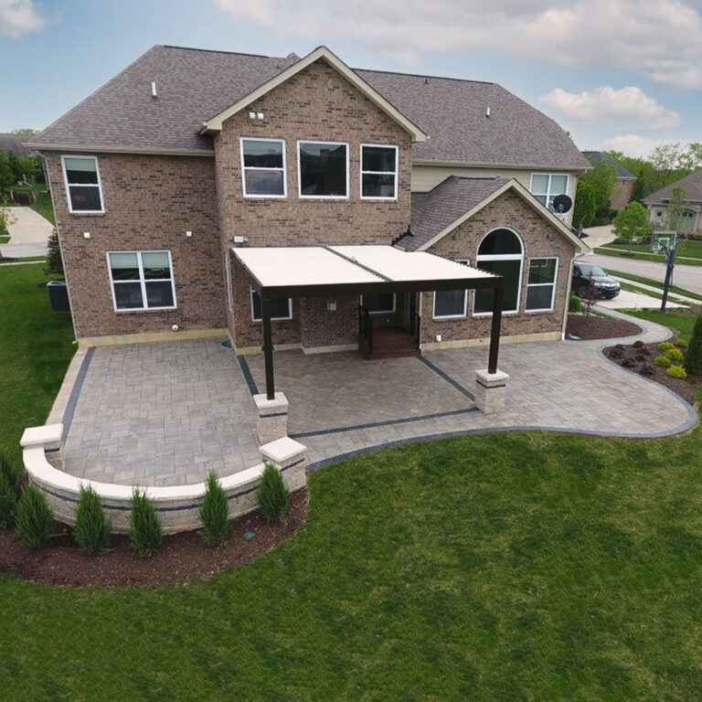 Aerial view of a dark-framed aluminum louvered pergola with closed cream louvers attached to a two-story brick home over a large curved paver patio with stone pier bases and a landscaped retaining wall