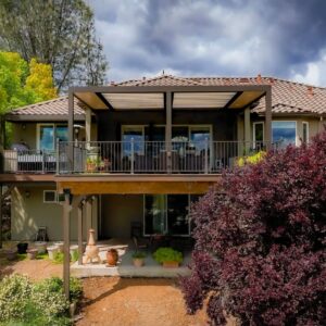Dark aluminum louvered pergola attached to a tile-roofed hillside home above a two-level backyard with mature ornamental trees and a chiminea below