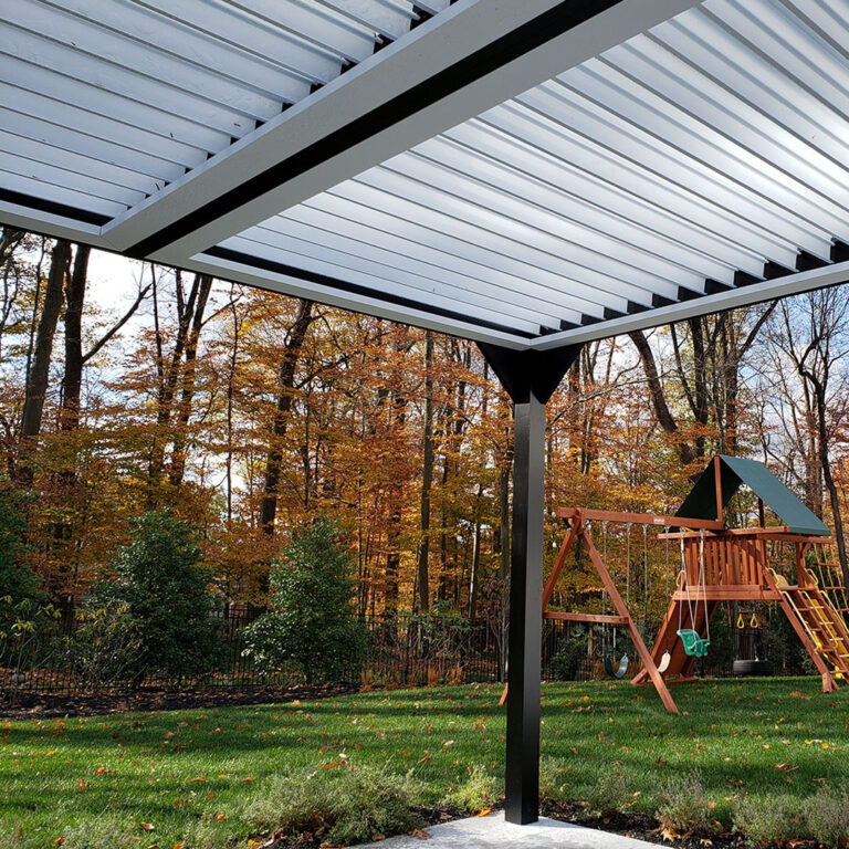Under-canopy view of a dark-framed aluminum pergola with fully open white louvers overlooking a backyard lawn with a wooden playset and autumn foliage