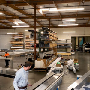 Workers handling aluminum louvered pergola components at assembly stations inside a large U.S. fabrication facility with structural extrusions stacked throughout.